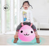 A young girl is holding a pink inflatable hopper ball with a face design on her lap in a bright room.