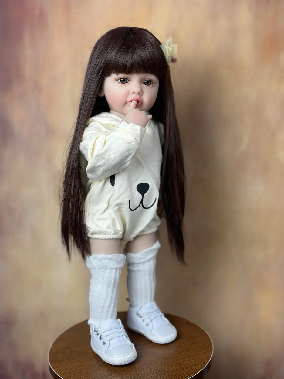 A lifelike 55 cm soft silicone baby doll with long dark hair wearing a white outfit and knee socks.
