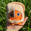 An orange Pochita plush doll with a light beige head covering is held in a hand against a green grassy background.