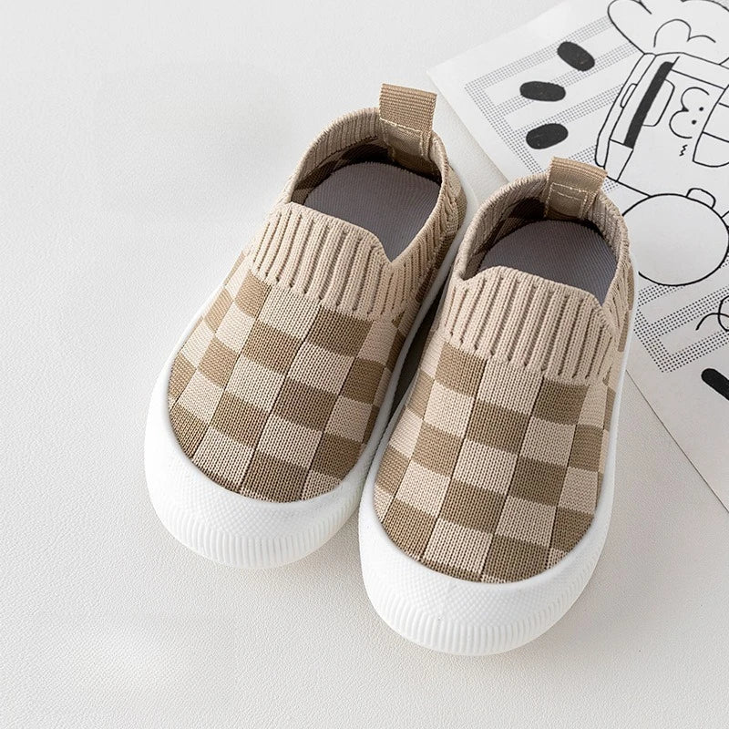 A pair of beige and tan plaid slip-on toddler shoes with knitted texture resting on a white surface next to a piece of paper.