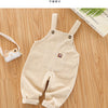 A beige baby cotton overall with adjustable straps lying on a wooden surface.