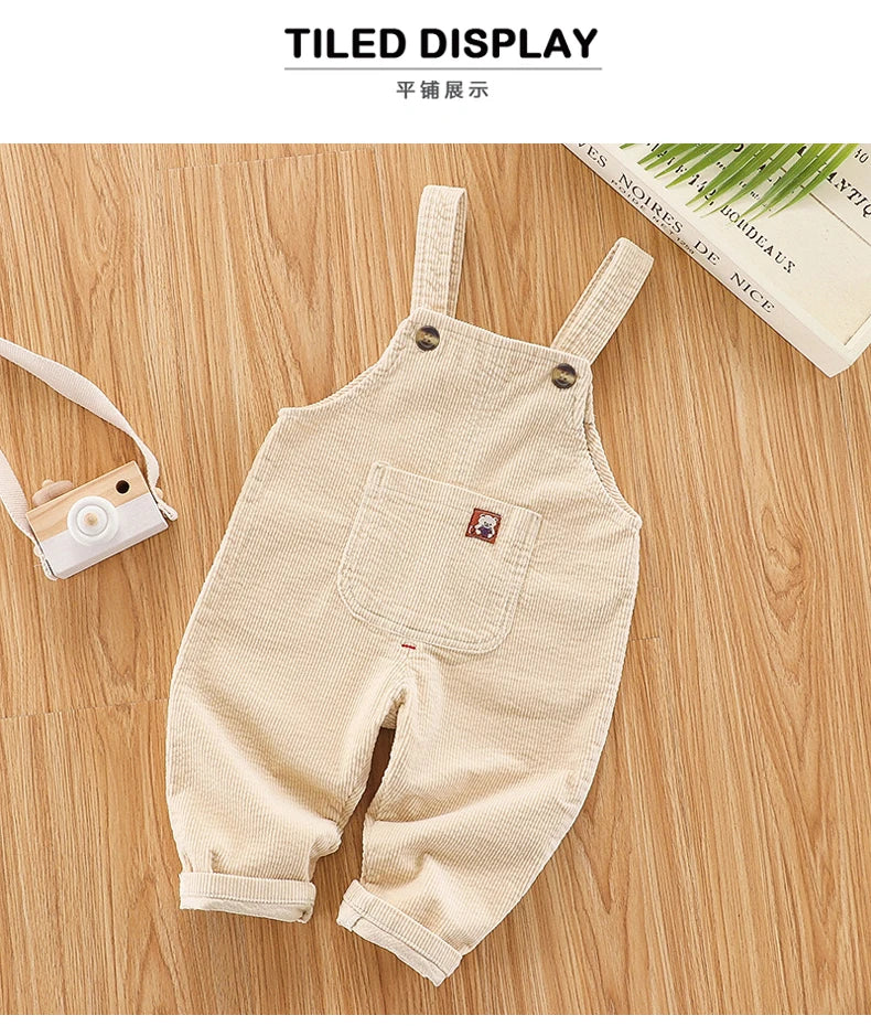 A beige baby cotton overall with adjustable straps lying on a wooden surface.