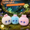 Three fluffy pink and blue rabbit plush toys with glowing eyes set against a lush green forest background.