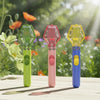 Three handheld insect catchers in green, pink, and yellow with blue grips resting on a wooden surface outdoors.