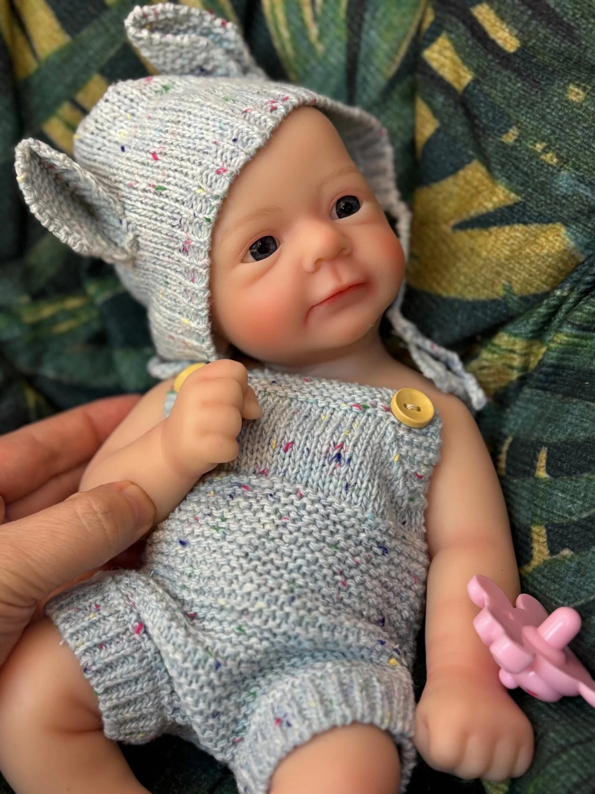 A hand-painted silicone reborn doll wearing a knitted hat and outfit, resting on green fabric.