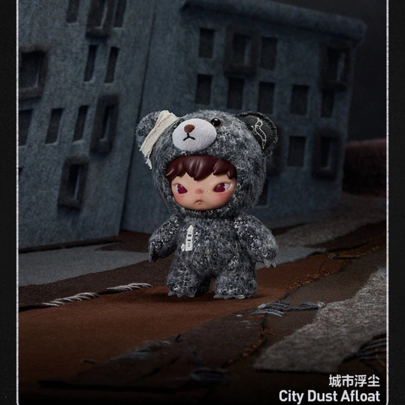 A small, dark gray bear plush doll with brown hair and a white ear covering sitting on a brown surface.