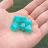 A hand holds a set of seven iridescent teal acrylic polyhedral dice on a green background.