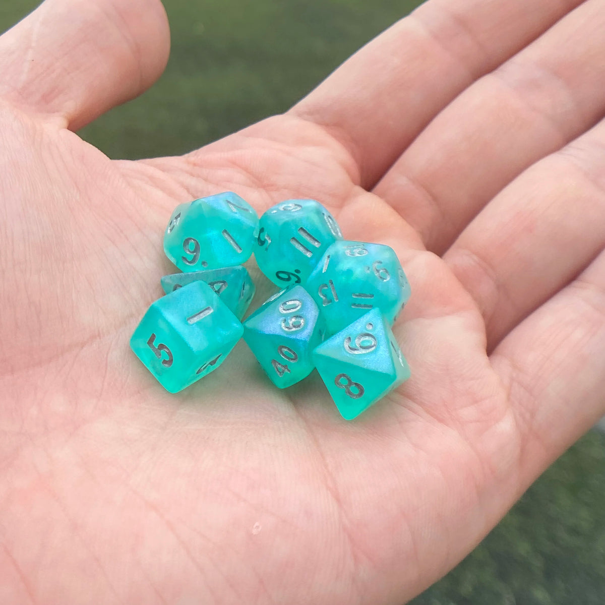 A hand holds a set of seven iridescent teal acrylic polyhedral dice on a green background.