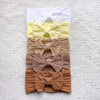A stack of five beige and yellow fabric bow baby headbands arranged vertically on a white textured background.