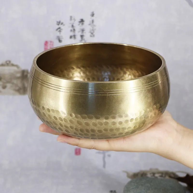 A hand holds a brass singing bowl with a mallet, used for meditation and sound therapy.
