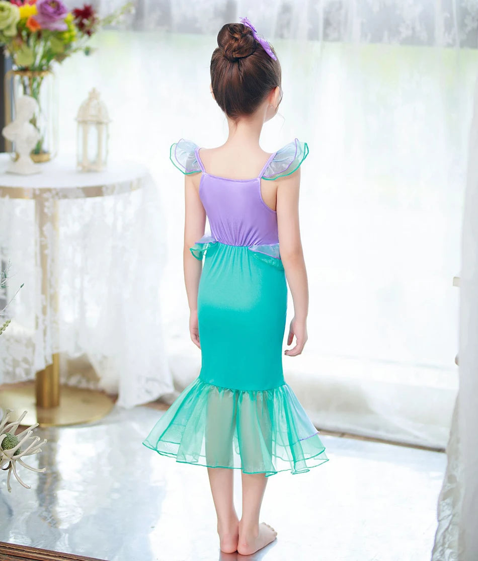 A girl models a purple and teal mermaid fantasy dress with ruffled fishtail skirt against a bright window background.
