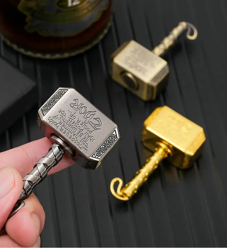 A hand holds a silver hammer-shaped metal fidget spinner with a gold handle and a brass-colored component on a dark surface.