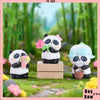 A white and black panda collectible figure with pink accents is displayed on a wooden block in a lush green outdoor setting.