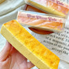 A yellow butter stick squishy toy is held in a hand, next to a packaged cheese cake product.