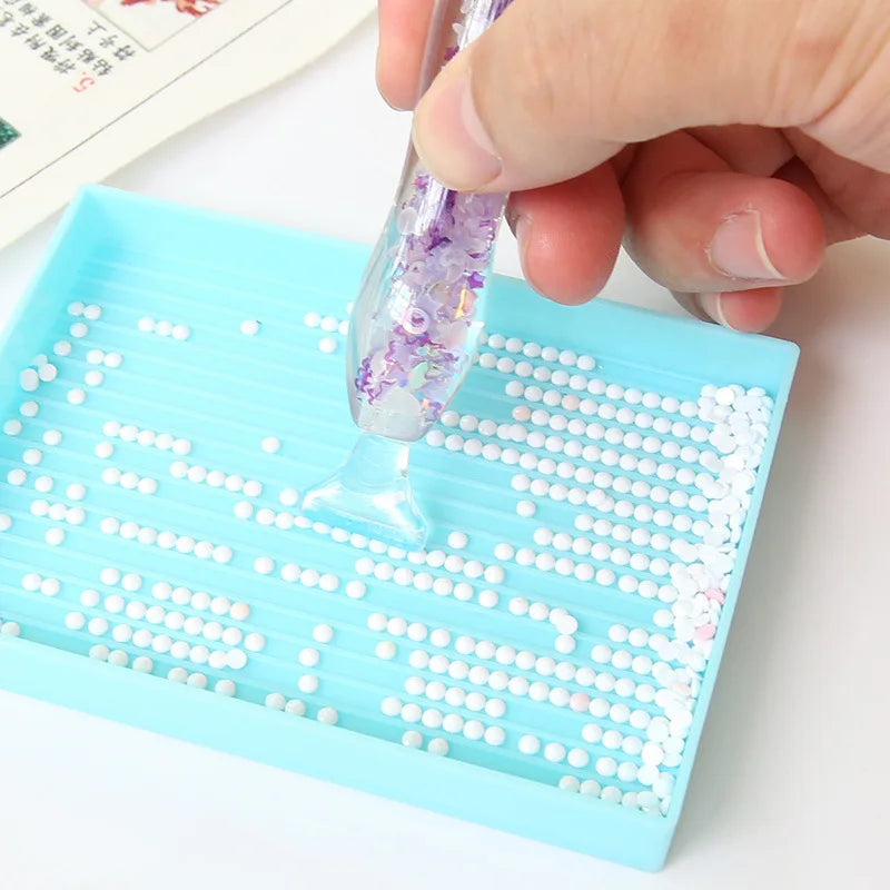 A hand is dipping a glow-in-the-dark diamond painting pen into a tray of small white and purple beads.