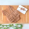 A brown lace baby romper with ruffled details and a matching brown bow headband displayed on a wooden board.