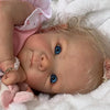 A close-up photo of a lifelike reborn baby doll with light blonde hair and blue eyes lying on white fluffy fabric.