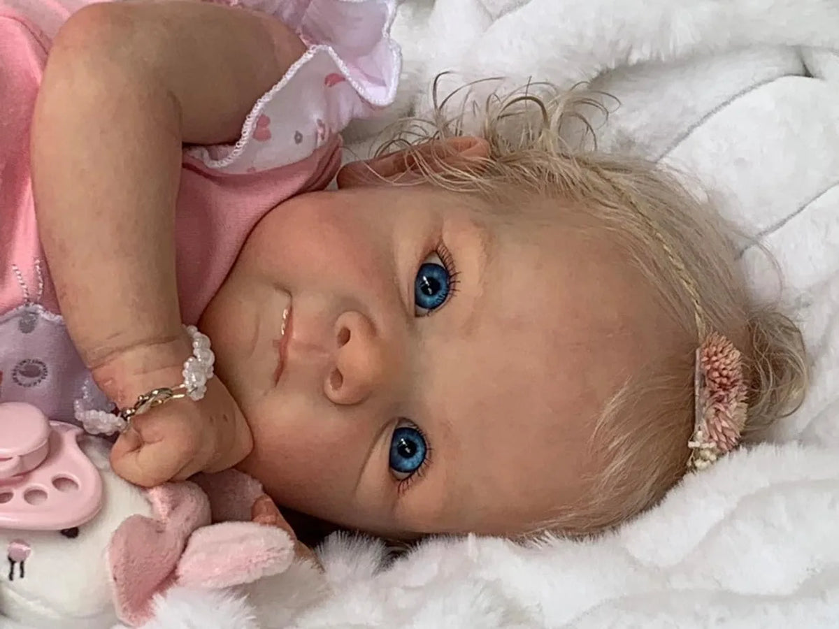 A close-up photo of a lifelike reborn baby doll with light blonde hair and blue eyes lying on white fluffy fabric.
