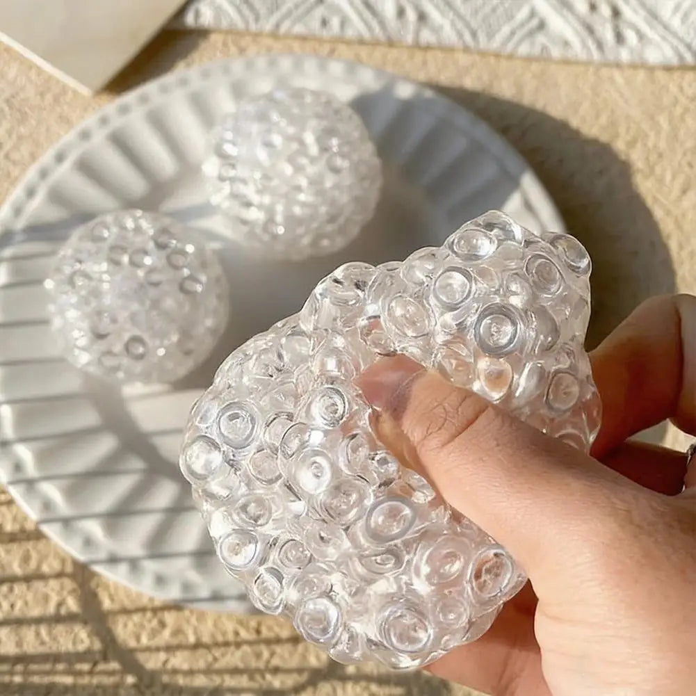 A hand is squeezing a clear, textured squishy bead stress ball on a white plate.