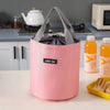 A pink stackable bento lunch bag with gray fabric handles sits on a white table next to juice bottles.