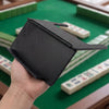 A hand holds a black foldable mahjong card holder with a non-slip stand on a green table surrounded by tiles.