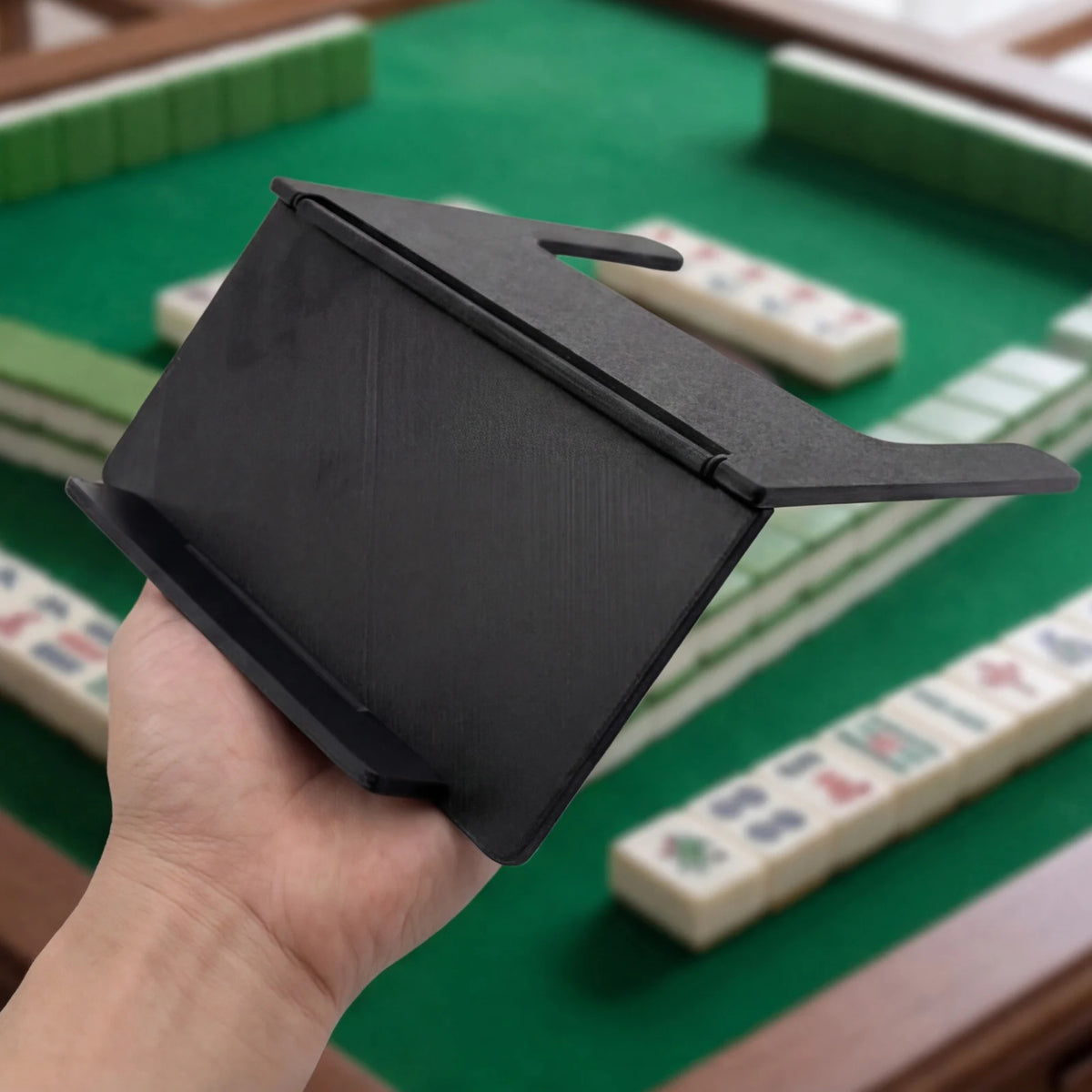 A hand holds a black foldable mahjong card holder with a non-slip stand on a green table surrounded by tiles.