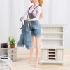 A mini doll wearing a denim overalls set and white t-shirt standing in front of a white wooden cabinet.