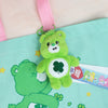 A green Care Bears plush keychain charm is attached to a pink strap against a teal fabric background.