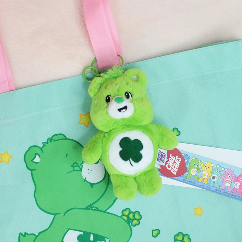 A green Care Bears plush keychain charm is attached to a pink strap against a teal fabric background.
