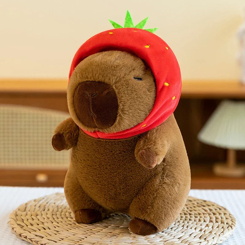 A brown capybara plush toy wearing a red hood and topped with a green leaf, sitting on a woven mat.