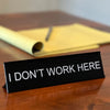 A black desk sign with white text that reads I DON'T WORK HERE is placed on a wooden surface.