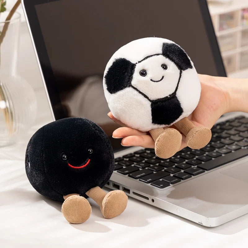 A hand holds a white and black plush sports ball companion with tan feet next to a laptop.