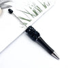 A black ballpoint pen with a diamond sequin bead section is shown next to some green grass and white paper.