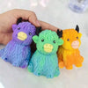 Three squishy toys shaped like Highland cows in purple, green, and orange colors being held by a hand.