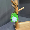 A handmade crochet frog charm with green and white yarn, attached to a gold-colored key ring hanging from a wooden stick.