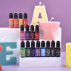 A set of sixteen liquid candle wax pigment colors in glass bottles arranged around a large letter A and an E.