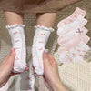 A pair of white floral cotton kids socks is being held up against a light beige background.