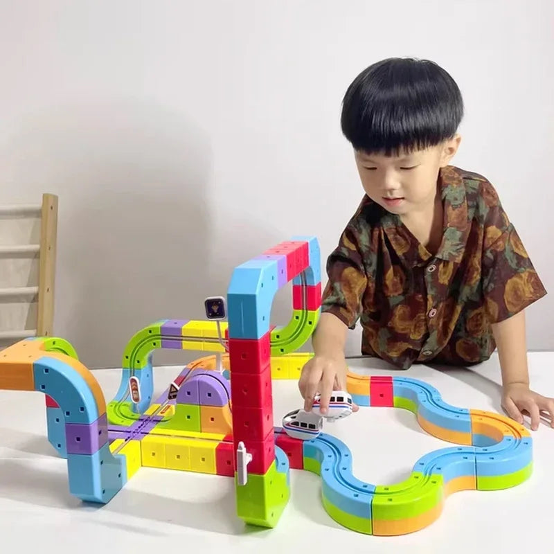 A young boy is building a colorful track train set using wooden and plastic blocks on a white table.