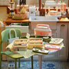 A miniature doll picnic lunch box with a handle is displayed on a table in front of a kitchen window.