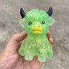 A hand holds a bright green glitter Highland cow squishy stress relief toy with black horns.