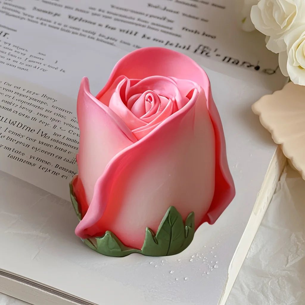 A pink rose silicone candle mold with green leaves resting on a white surface next to a piece of paper.