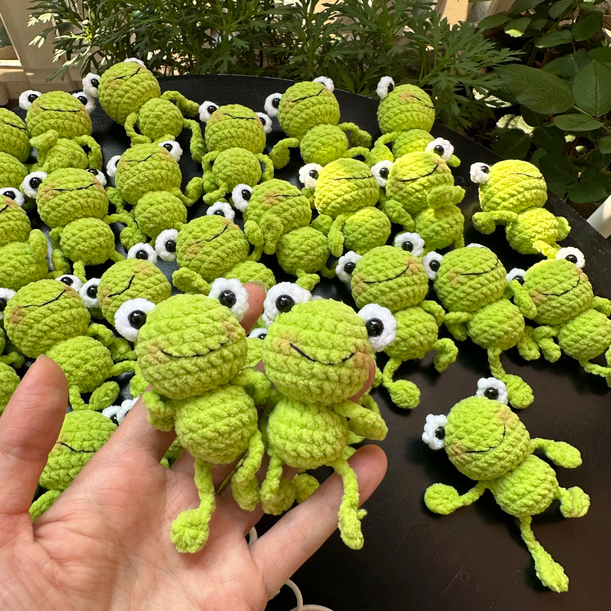 A hand holds a collection of handmade crochet frog keychains in various shades of green and yellow.