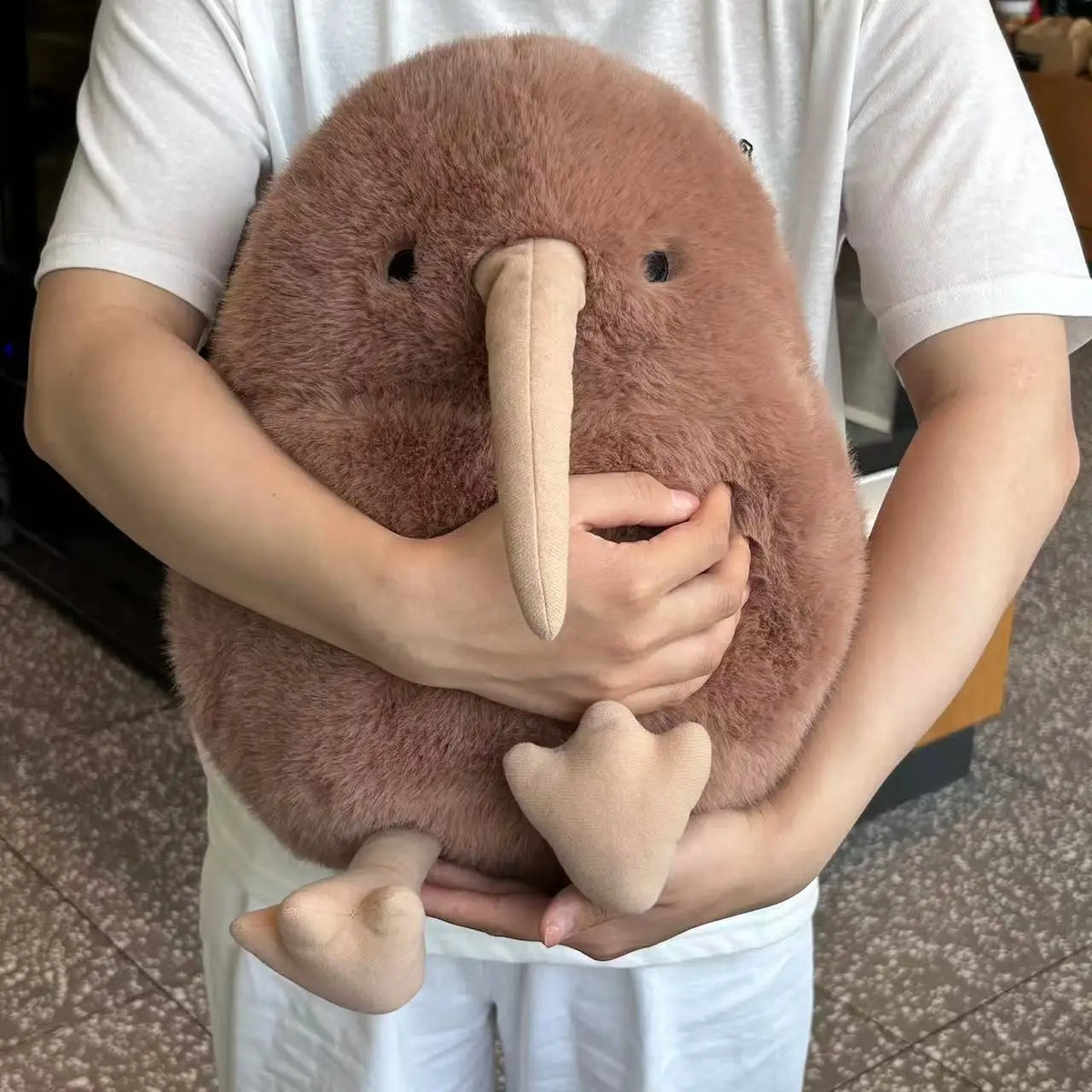 A person is holding an oversized brown kiwi bird plush toy with a long beak and soft feathers.