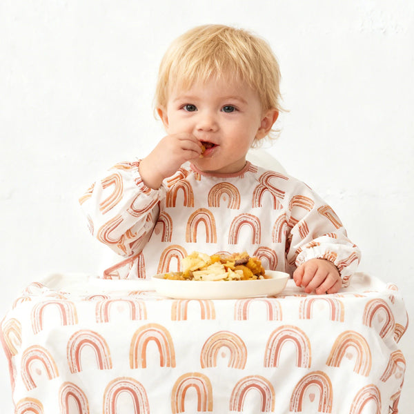A baby is eating food while wearing a long-sleeved waterproof meal bib with a rainbow pattern.