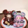 A plush charm of a bear character with brown fur and pink accents displayed on a round plate.