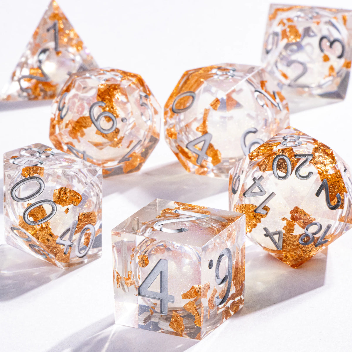 A collection of polyhedral dice with a clear, galaxy finish and gold accents are displayed on a white surface.