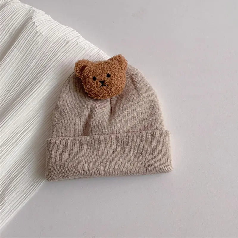 A knitted baby hat in beige with a brown bear ear patch resting on a white background.
