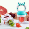 A silicone baby food feeder with a teether is shown with various fruits and vegetables on a white plate.