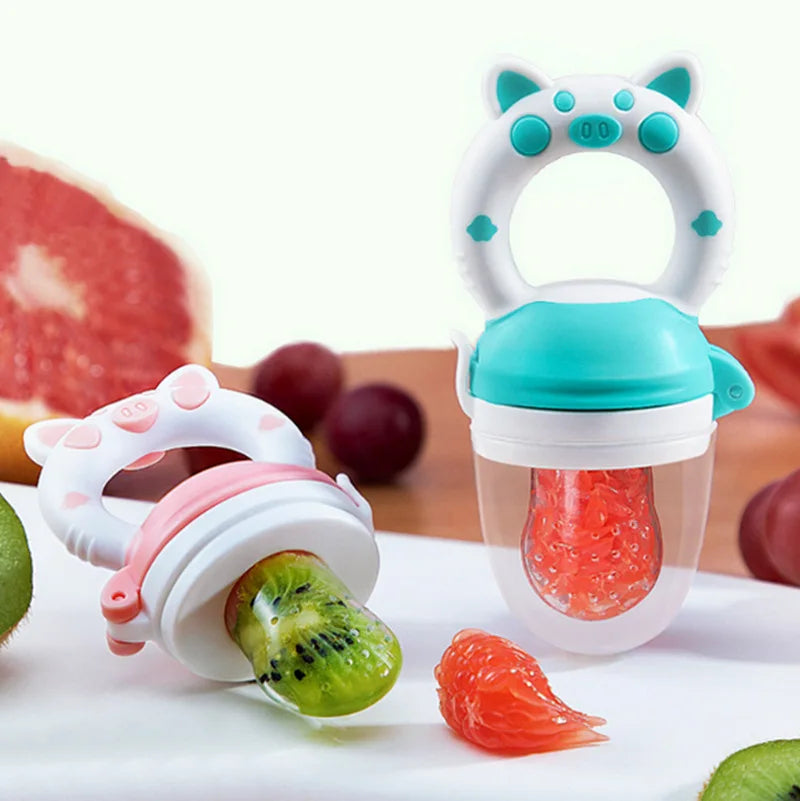 A silicone baby food feeder with a teether is shown with various fruits and vegetables on a white plate.