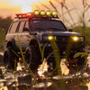 A black 1:18 scale remote control off-road climbing car with bright headlights driving through wet foliage at sunset.